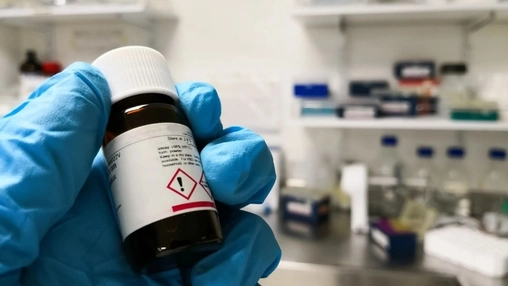 Gloved hand holding a labeled chemical sample vial in a laboratory