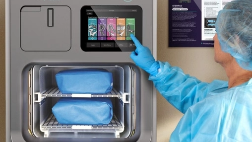 Healthcare worker in blue scrubs and gloves operating a STERRAD steriliser touchscreen with wrapped packs